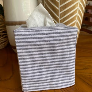 May include: A fabric tissue box cover with a gray and white striped pattern. White tissues are visible extending from the top opening. The box is sitting on a wooden surface, with decorative vases in the background.