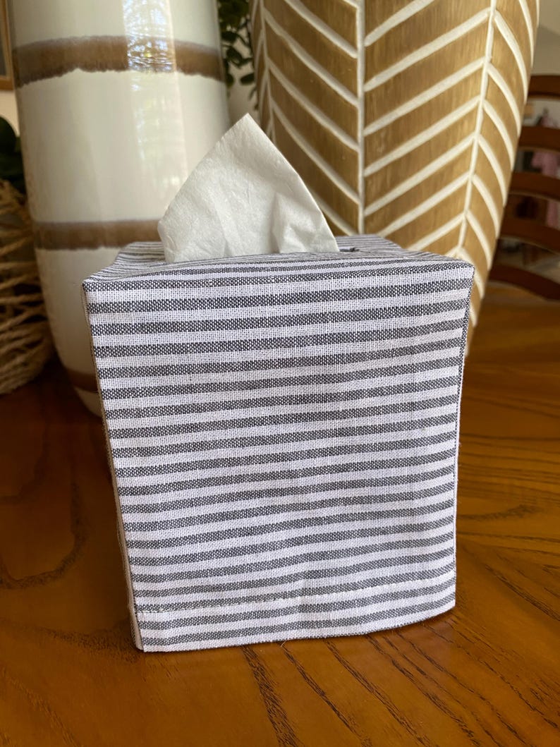 May include: A fabric tissue box cover with a gray and white striped pattern. White tissues are visible extending from the top opening. The box is sitting on a wooden surface, with decorative vases in the background.