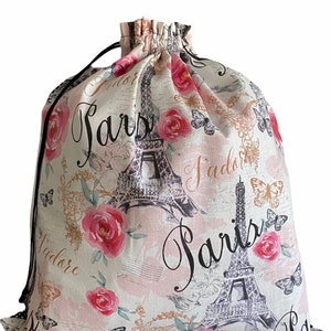 May include: A drawstring bag with a Parisian theme. The bag features a cream-colored background with pink roses, butterflies, and the Eiffel Tower. The word "Paris" is printed in a script font. A black drawstring is visible at the top.