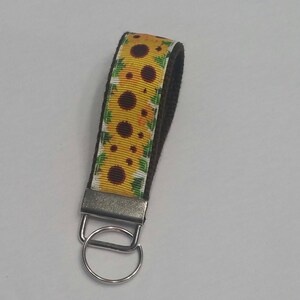 May include: A sunflower wristlet keychain with a yellow and brown floral pattern. The wrist strap is a dark brown color, and the keychain has a silver metal clasp and ring.