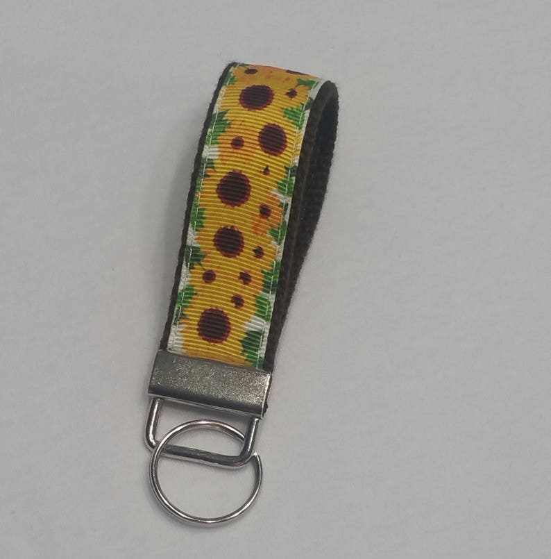 May include: A sunflower wristlet keychain with a yellow and brown floral pattern. The wrist strap is a dark brown color, and the keychain has a silver metal clasp and ring.
