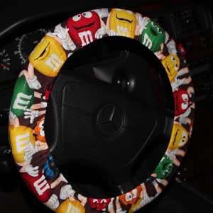 May include: A colorful steering wheel cover featuring a repeating pattern of M&M's candies in various colors. The cover is wrapped around a black steering wheel with a Mercedes-Benz logo.