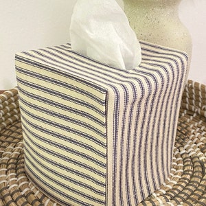 May include: A square tissue box cover with a cream and navy blue striped pattern. White tissues are pulled from the top. The box is sitting on a woven tray, with a decorative vase and dried flowers in the background.