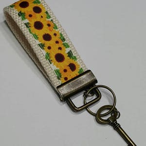 May include: A beige fabric keychain wristlet with a yellow sunflower pattern and a bronze-colored metal clasp and key. The key has a vintage design. The sunflower pattern has yellow petals and brown centers.