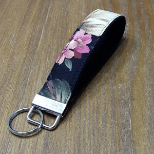 May include: A fabric key fob with a floral pattern in pink, green, and beige on a black background. The key fob has a silver metal clasp and a split ring for attaching keys. The key fob is approximately 6 inches long.