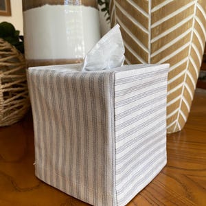 May include: A square tissue box cover with a white and gray striped pattern. The box is made of fabric and has a tissue sticking out of the top. The cover is sitting on a wooden surface.