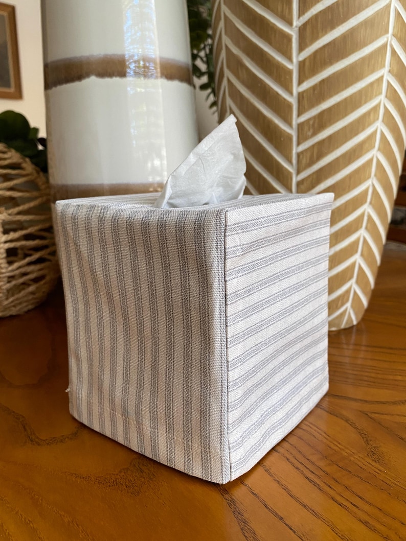 May include: A square tissue box cover with a white and gray striped pattern. The box is made of fabric and has a tissue sticking out of the top. The cover is sitting on a wooden surface.