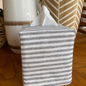 May include: A square tissue box cover with a black and white striped pattern. White tissues are visible at the top. The cover is made of fabric and sits on a wooden surface. Decorative vases are in the background.