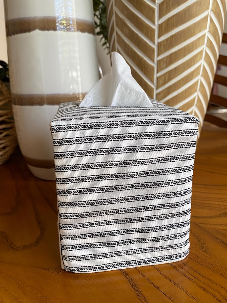 May include: A square tissue box cover with a black and white striped pattern. White tissues are visible at the top. The cover is made of fabric and sits on a wooden surface. Decorative vases are in the background.