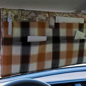 May include: A car visor organizer with multiple pockets. The organizer is made of fabric with a plaid pattern in shades of brown, black, and white. The top section features a map design. A pen and several papers are visible in the pockets.