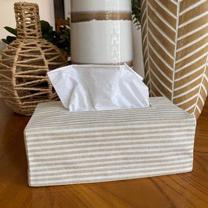 May include: A rectangular tissue box cover with a beige and white striped pattern. White tissues are visible at the top. The box is on a wooden surface, with decorative vases in the background.
