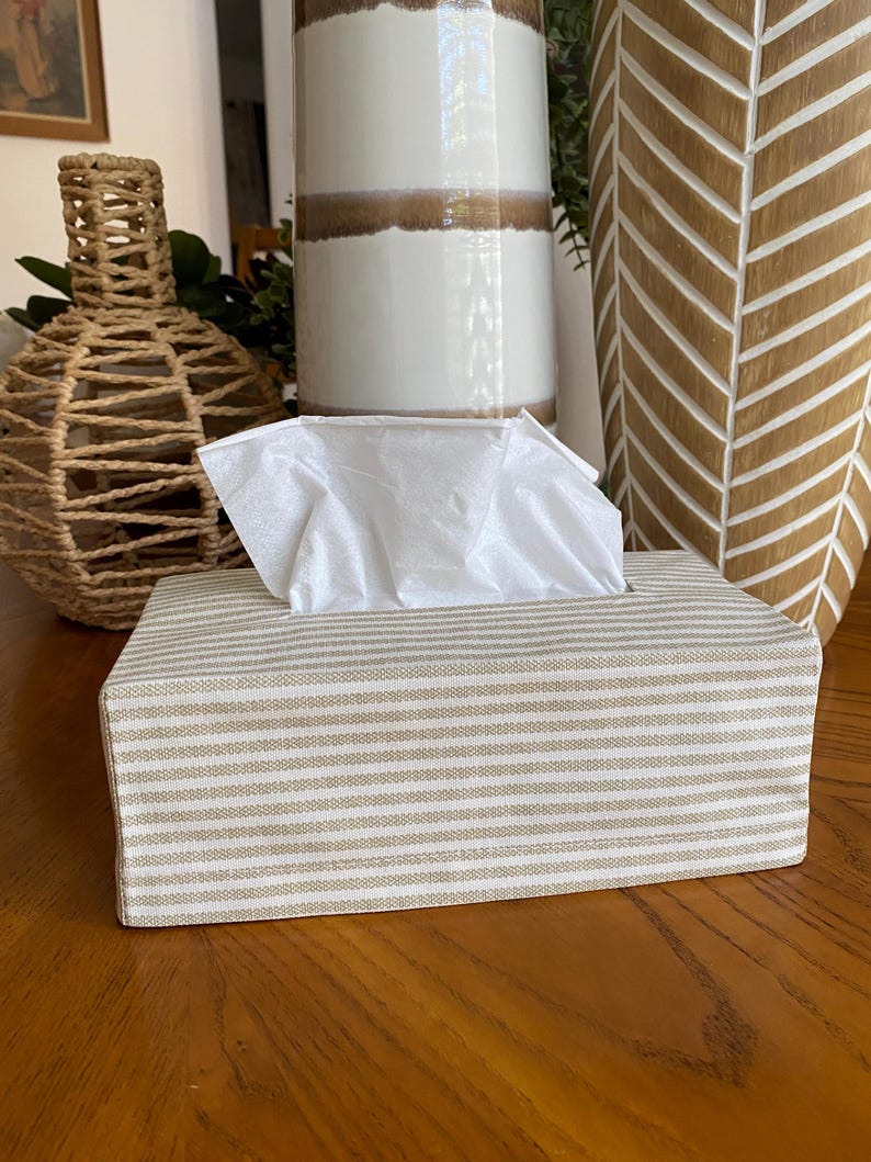 May include: A rectangular tissue box cover with a beige and white striped pattern. White tissues are visible at the top. The box is on a wooden surface, with decorative vases in the background.