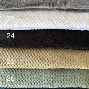 Op de afbeelding: Een stapel van zes gevouwen handdoeken met verschillende texturen en kleuren, genummerd van 22 tot 27. De handdoeken zijn grijs, wit, zwart, beige, saliegroen en donker marineblauw en hebben elk een duidelijk zichtbaar weefpatroon.