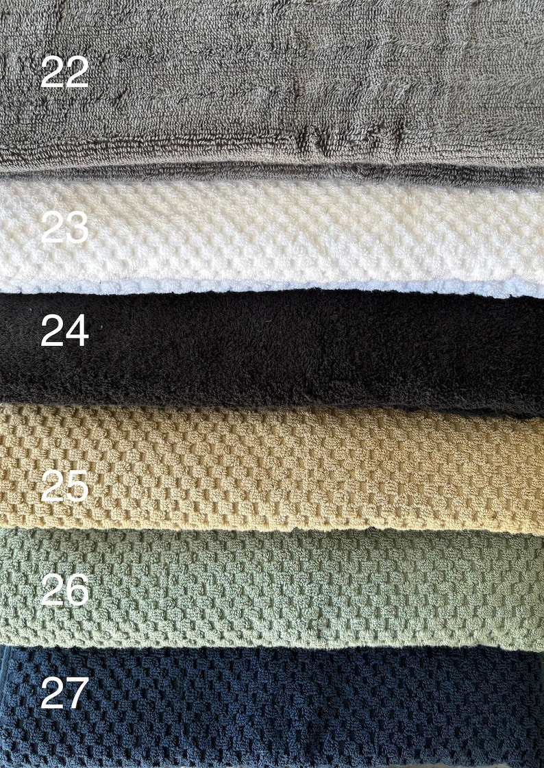 Op de afbeelding: Een stapel van zes gevouwen handdoeken met verschillende texturen en kleuren, genummerd van 22 tot 27. De handdoeken zijn grijs, wit, zwart, beige, saliegroen en donker marineblauw en hebben elk een duidelijk zichtbaar weefpatroon.