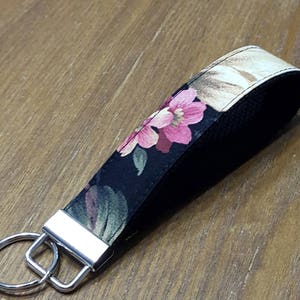 May include: A floral key fob with a black background and pink flowers. The key fob has a silver metal key ring and a loop for the wrist. The fabric has a floral pattern with pink, green, and tan colors.