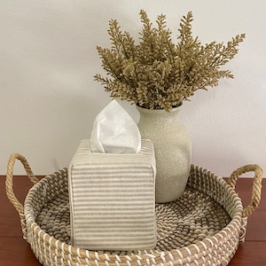 May include: A decorative woven tray with handles holds a tissue box and a vase with dried plants. The tissue box is covered in a beige and white striped fabric. The vase is a light beige color.