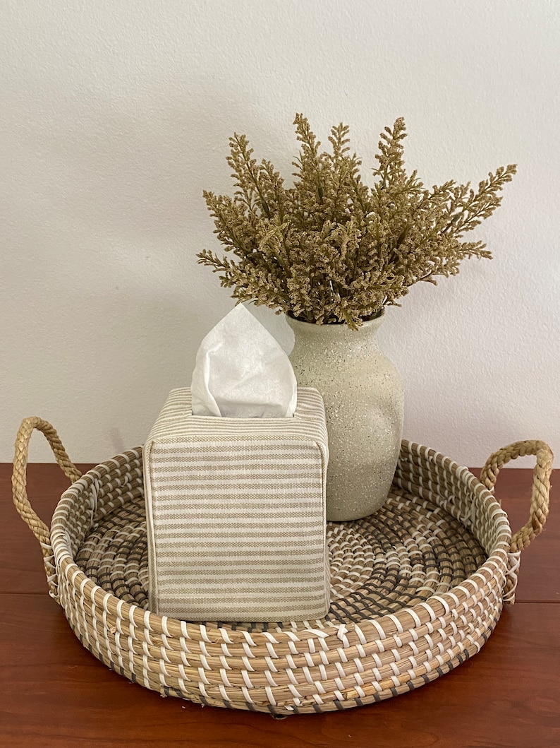 May include: A decorative woven tray with handles holds a tissue box and a vase with dried plants. The tissue box is covered in a beige and white striped fabric. The vase is a light beige color.
