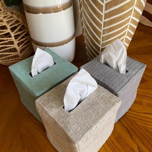 May include: Three decorative tissue box covers in shades of green, beige, and gray. Each box is rectangular with a textured fabric exterior and a rectangular opening for tissue dispensing. White tissues are visible.