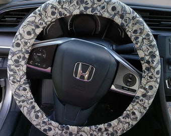 Puede incluir: Una funda para volante con un patrón repetido de calaveras grises sobre un fondo beige claro. La funda está envuelta alrededor de un volante negro con un emblema plateado de Honda en el centro. El salpicadero del coche es visible en el fondo.