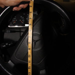 May include: A black steering wheel with a Mercedes-Benz logo is measured with a ruler. The ruler shows a measurement of approximately 16 inches. The steering wheel is in a car interior.