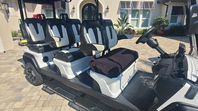 Op de afbeelding: Een witte golfkar voor zes personen met zwart-wit gestoffeerde stoelen, een zwart stuur en een zwart dak. Het voertuig is voorzien van zijarmsteunen, een zwarte vloermat en wielen met meerdere spaken, ontworpen voor buitenvervoer.