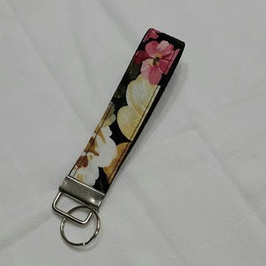 May include: A floral key fob with a black background and a pattern of pink, yellow, and white flowers. The key fob has a silver-colored metal ring and a rectangular metal piece.