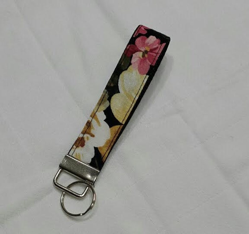 May include: A floral key fob with a black background and a pattern of pink, yellow, and white flowers. The key fob has a silver-colored metal ring and a rectangular metal piece.
