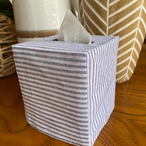May include: A square tissue box cover with a gray and white striped pattern. A white tissue is partially visible at the top. The box is made of fabric and sits on a wooden surface. Decorative vases are in the background.