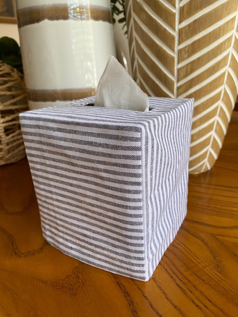 May include: A square tissue box cover with a gray and white striped pattern. A white tissue is partially visible at the top. The box is made of fabric and sits on a wooden surface. Decorative vases are in the background.