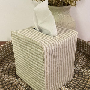 May include: A square tissue box cover with a beige and white striped pattern. The cover is made of fabric and sits on a woven tray. White tissues are visible, emerging from the top opening. A vase with dried flowers is in the background.