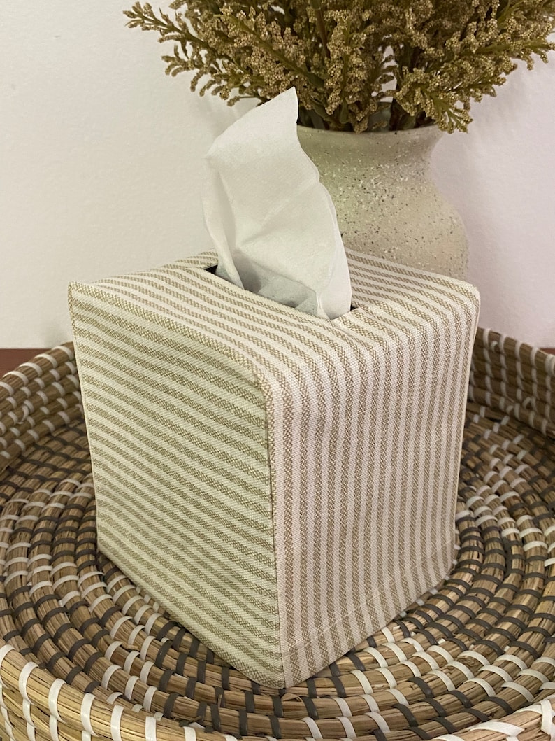 May include: A square tissue box cover with a beige and white striped pattern. The cover is made of fabric and sits on a woven tray. White tissues are visible, emerging from the top opening. A vase with dried flowers is in the background.