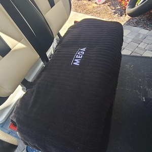 Op de afbeelding: Een zwarte rechthoekige stoelhoes met een geribbelde textuur en het woord MEGA geborduurd in wit garen, geplaatst over de bank van een voertuig met cr&egrave;mekleurige en zwarte bekleding. De hoes heeft een horizontale ori&euml;ntatie.
