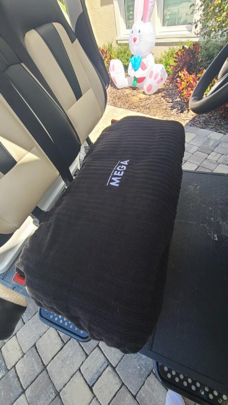 Op de afbeelding: Een zwarte rechthoekige stoelhoes met een geribbelde textuur en het woord MEGA geborduurd in wit garen, geplaatst over de bank van een voertuig met cr&egrave;mekleurige en zwarte bekleding. De hoes heeft een horizontale ori&euml;ntatie.