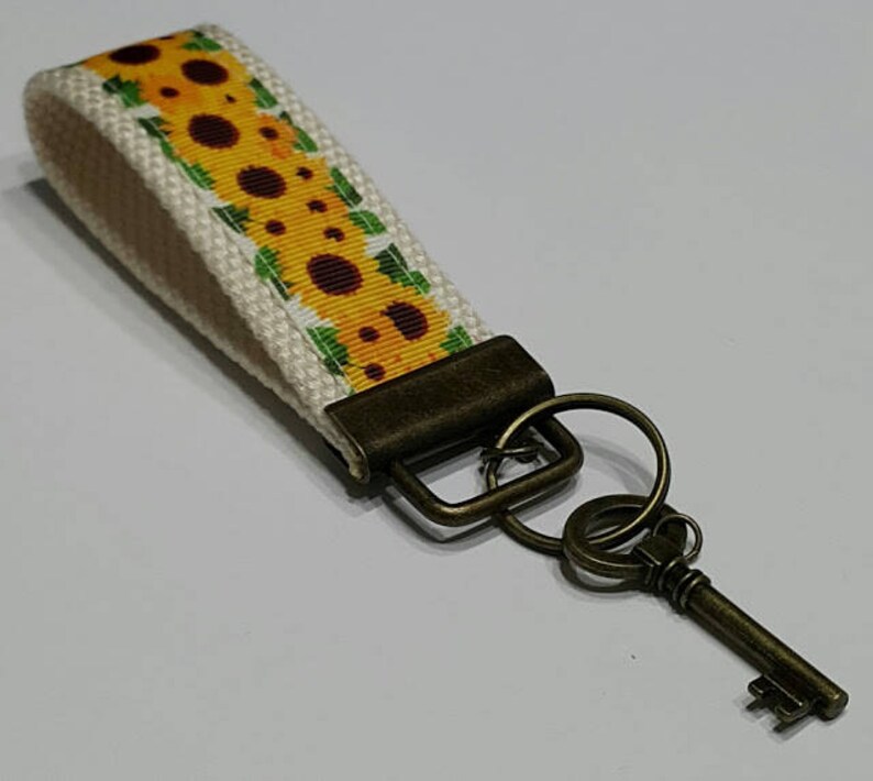 May include: A key fob with a sunflower pattern on a cream-colored background. The key fob is attached to a bronze-colored metal key and hardware. The key is a vintage style.