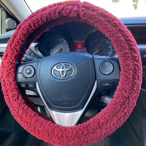 May include: A burgundy fuzzy steering wheel cover on a black steering wheel. The cover is made of a soft, plush material and is wrapped around the steering wheel. The car's dashboard and Toyota logo are visible.