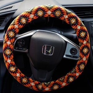 May include: A steering wheel cover with a vibrant geometric pattern in shades of orange, yellow, black, and white. The cover is wrapped around a black steering wheel, enhancing the car's interior with a touch of color and design.
