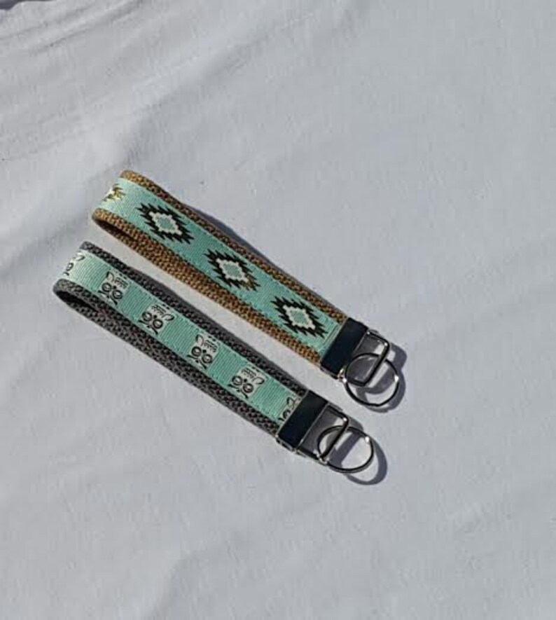 May include: Two wrist keychains with woven fabric straps. One is gray with a repeating owl pattern, the other is tan with a turquoise and black geometric design. Both have silver-tone metal hardware.