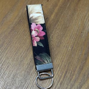 May include: A floral key fob with a black background, featuring pink and white flowers and green leaves. The fabric strap is attached to a silver-colored metal ring and hardware.