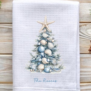 May include: White kitchen towel with a blue and white seashell Christmas tree design. The tree is topped with a starfish and the towel is personalized with the text "The Reeces".