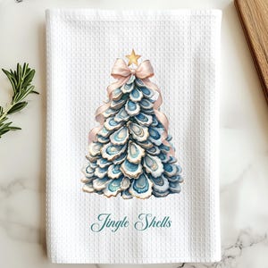 May include: White waffle-weave kitchen towel featuring a watercolor Christmas tree design made of blue and white oyster shells. A gold star tops the tree, with a pink ribbon bow. The words "Jingle Shells" are written in script below the tree.