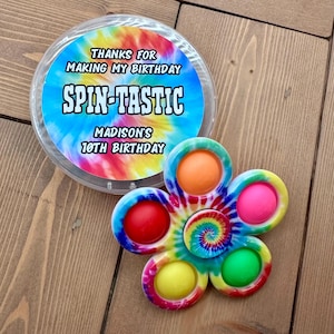 May include: A tie-dye flower-shaped fidget spinner with six colored bubbles and a central tie-dye design. A clear plastic container with the text "THANKS FOR MAKING MY BIRTHDAY SPIN-TASTIC MADISON'S 10TH BIRTHDAY" is next to the spinner.