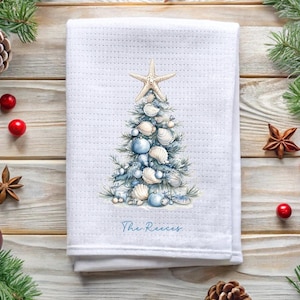 May include: A white waffle-weave kitchen towel featuring a printed illustration of a Christmas tree decorated with various seashells and light blue ornaments, topped with a starfish. The text "The Reeces" is printed in blue below the tree.