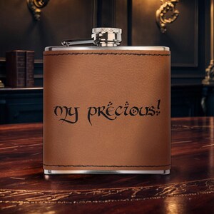 May include: A brown leather-wrapped flask with a silver cap. The flask has the phrase "my precious!" in black script. The flask is rectangular and sits on a wooden table. The background includes a dark room with bookshelves and lamps.