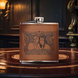 Peut inclure: Une flasque recouverte de cuir marron avec un dessus argenté et un motif gravé décoratif d'une femme avec une fleur dans les cheveux. La flasque est posée sur une table en bois.