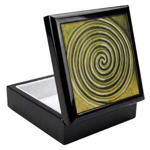 May include: A square black keepsake box with a hinged lid featuring a centered olive green and black spiral pattern. The interior of the box is lined with a white fabric material, and the exterior has a smooth, glossy finish on all sides.