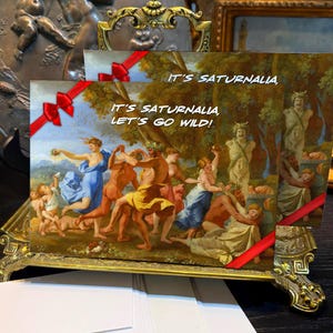 Puede incluir: Tarjetas de felicitación con una pintura clásica de una escena festiva, sostenidas en un soporte decorativo de color dorado. Las tarjetas están atadas con cintas rojas y presentan el texto "IT'S SATURNALIA, LET'S GO WILD!"