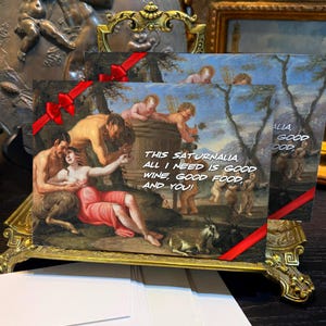 Puede incluir: Tarjetas de felicitación con una pintura de estilo renacentista, con figuras y el texto "THIS SATURNALIA, ALL I NEED IS GOOD WINE, GOOD FOOD, AND YOU!" Las tarjetas se muestran en un soporte dorado decorativo con una cinta roja.