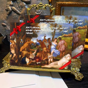 Puede incluir: Tarjetas de felicitación con una escena de pintura renacentista, sostenidas en un soporte dorado decorativo. Las tarjetas tienen acentos de cinta roja y el texto dice "¡SATURNALIA! ES EL MOMENTO MÁS MARAVILLOSO DEL VINO DEL AÑO."