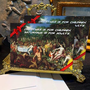Puede incluir: Tarjetas de felicitación con una escena de pintura clásica, que presenta una reunión festiva. Las tarjetas están adornadas con cintas rojas y el texto "Christmas is for children, Saturnalia is for adults."