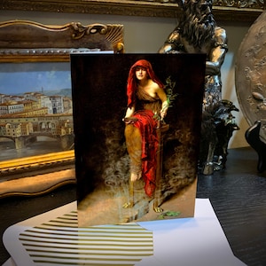 May include: Greeting card featuring a classical painting of a woman in a red hooded cloak, holding a branch. The artwork has a dark, dramatic aesthetic. The card is displayed on a striped surface, with other framed art pieces in the background.